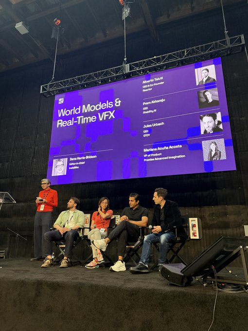 The image shows a group of five people sitting around a stage with a large screen behind them displaying information about "World Models & Real-Time VFX". The individuals are engaged in conversation, possibly discussing the content displayed on the screen or sharing their thoughts on the topic. The backdrop features a microphone and speaker system, suggesting that they might be part of an event or presentation related to the subject matter.