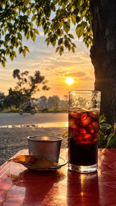 A tall glass of iced dark beverage and a small ceramic cup sit on a red table against a sunset backdrop with trees and a motorbike in the distance.