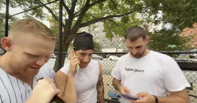 Three men stand together near a chain-link fence and tree, one looking at his phone while another talks on a cell phone; the man on the right wears a shirt reading 'type jawn'.