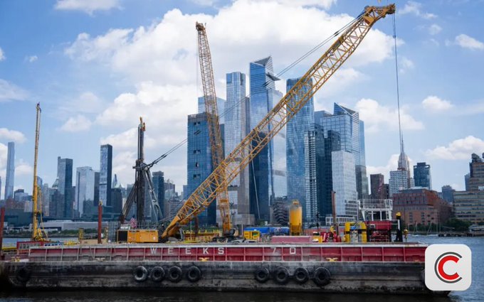 A construction barge with a yellow crane labeled 'WEEKS 730' on the water, set against a backdrop of the modern Manhattan skyline including Hudson Yards skyscrapers under a partly cloudy sky, with a 'C' logo in the bottom right corner.