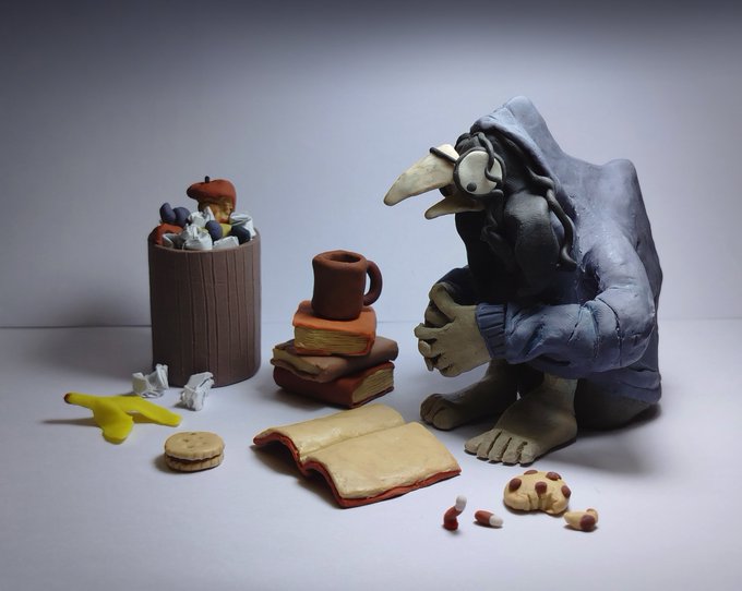 A claymation-style sculpture scene featuring a long-nosed, hooded character sitting beside a trash can filled with crumpled paper, books, a mug, cookies, and other small objects.