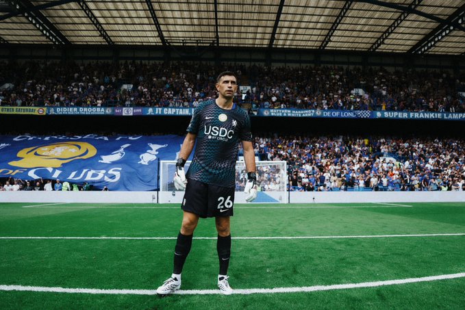 A soccer goalkeeper wearing a USDC-sponsored jersey with the number 26 stands on the pitch at a packed stadium, with Chelsea FC banners visible in the crowd. The image illustrates the brand's debut in professional sports sponsorship, showcasing prominent USDC logo placement on the player's kit.