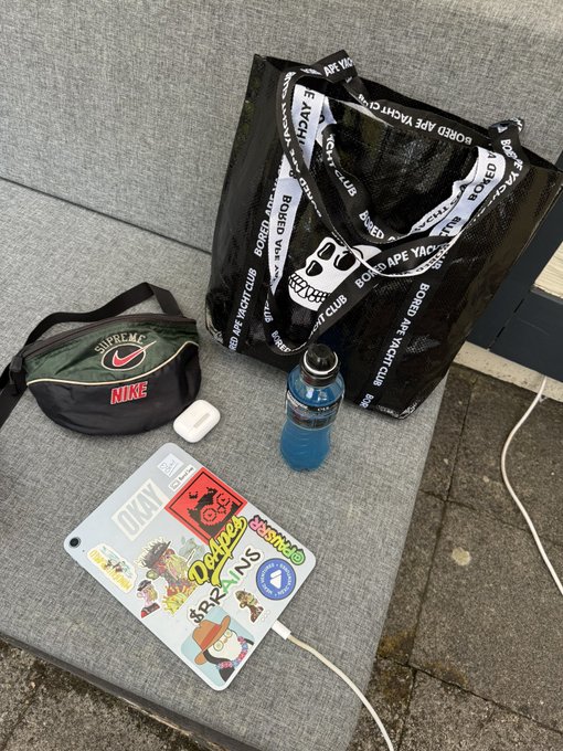 A flat lay of personal items on a gray cushion including a Supreme x Nike duffel bag, a Bored Ape Yacht Club tote, a blue water bottle, AirPods, and a sticker-covered laptop with a white cable.