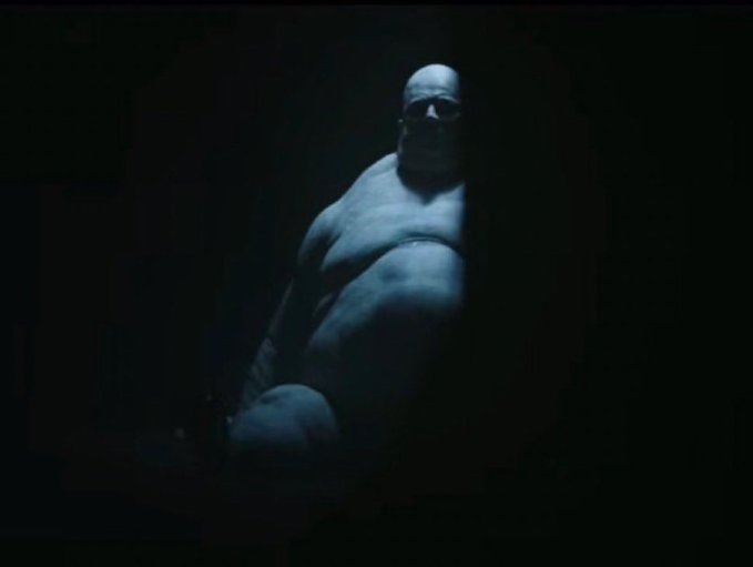 A dark, shadowy 3D render depicts a muscular humanoid figure with glowing sunglasses emerging from the blackness.