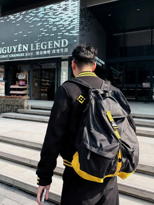 A person with short hair carries a black and yellow backpack while walking up steps outside a store with signage reading 'NGUYEN LEGEND' and 'THAT CHANGES LIFE'.