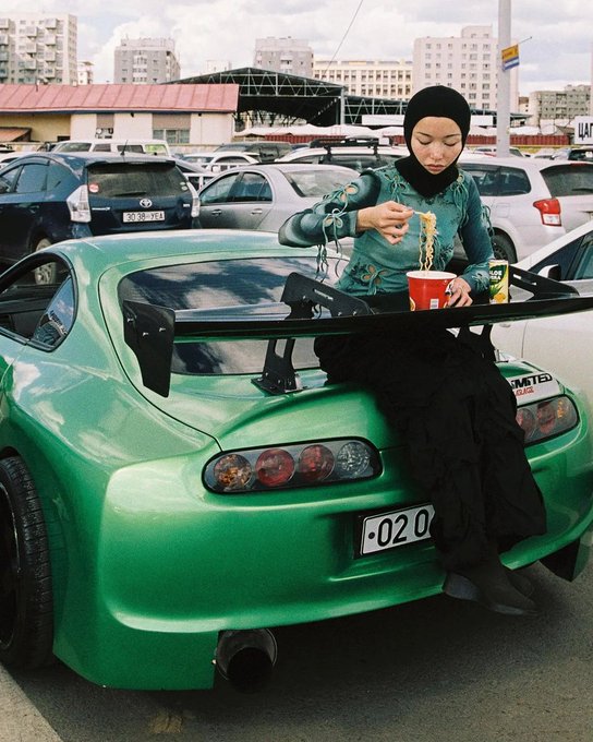A woman wearing a black hijab sits on the rear spoiler of a bright green sports car, eating noodles from a red cup in a parking lot.