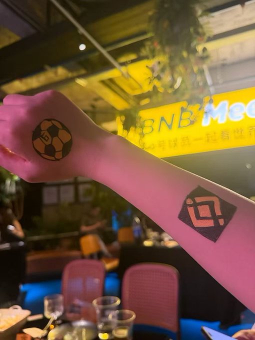 A person's arm is extended, showing a soccer ball stamp on the back of the hand and a 'BNB' logo stamp on the forearm, with a restaurant featuring a yellow 'BNB' sign in the background.