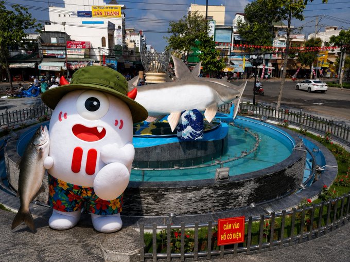 A cartoon mascot wearing a green safari hat and floral shorts poses next to a shark statue at a city roundabout, with a "CẤM VÀO HỒ CÓ ĐIỆN" warning sign visible. The mascot, appearing as a playful white character with vampire-like teeth, holds a fish, adding a whimsical contrast to the urban Vietnamese street scene that pairs with the post about trying one's luck with #TronBull.
