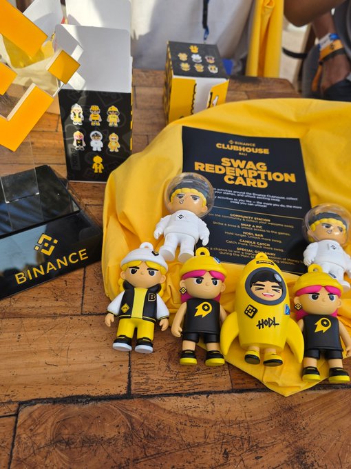 A collection of Binance Clubhouse Crew "Swag Redemption Card" figures and blind boxes arranged on a yellow shirt, featuring astronaut-themed mascots, with a black Binance-branded display card prominently visible on the left, fitting the Coinfest Day 2 giveaway mentioned in the post.