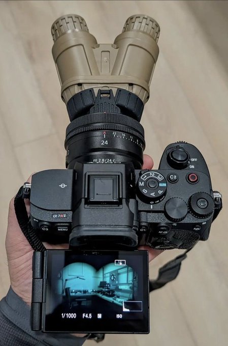 An overhead view of a Sony camera with an unusual setup of binoculars attached as a lens, held by a person; the flip-out screen displays a view of a room with settings of 1/1000, F4.5, and ISO visible.