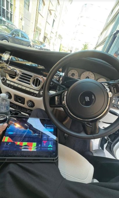 The image shows a person sitting inside a car with their legs crossed over each other while holding a tablet computer. The person appears to be using the tablet for some form of entertainment or communication. In the background, there are several cars parked on the street outside the vehicle.