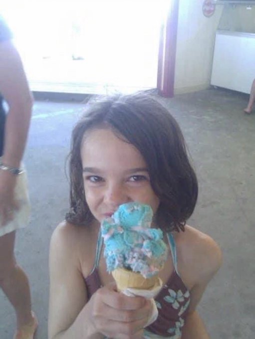 A young girl with shoulder-length brown hair smiles while eating a large cone of blue and orange ice cream.