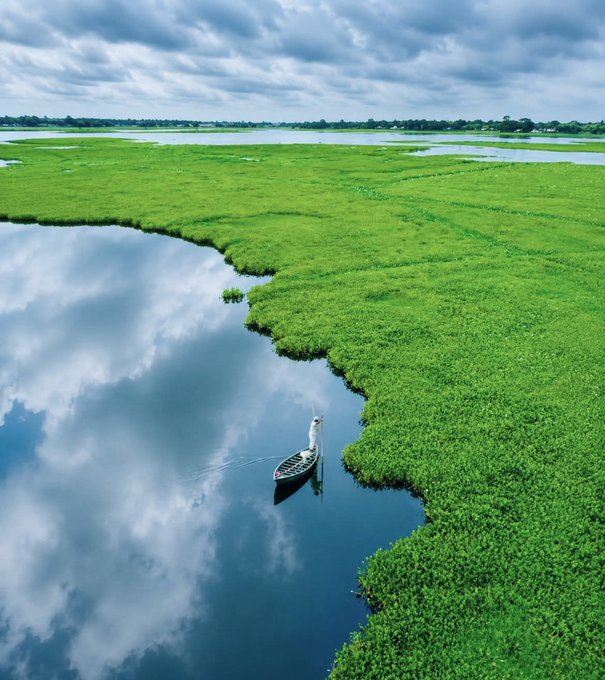 The image features a small boat floating on a body of water, surrounded by green grass. The sky above has clouds, suggesting that it might be early morning or late afternoon when the sun is low in the sky. The scene appears to be peaceful and serene, with no people visible around the boat.