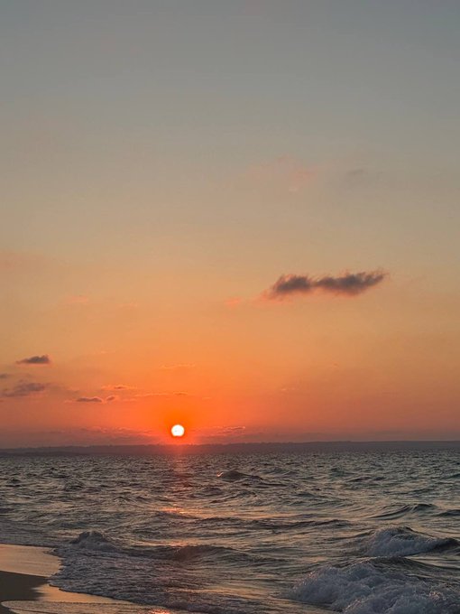 A scenic sunset over the ocean, with the sun low on the horizon, orange sky, and small waves reaching the sandy shore.