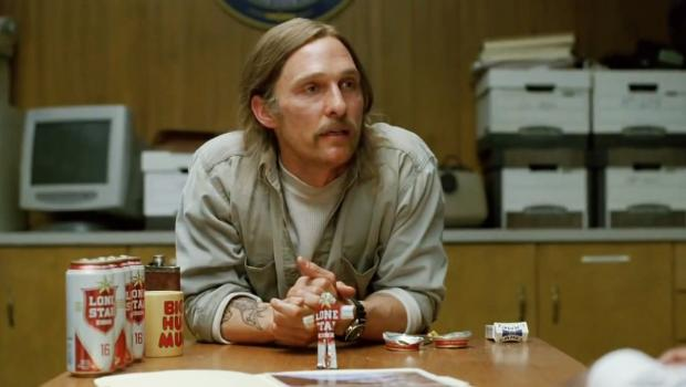 The image features an actor sitting at a desk with various objects, including several cans of beer scattered around him. The scene appears to be set in a room that also contains a TV monitor and a keyboard. A clock can be seen on one side of the room as well. The presence of these items suggests that this could be a casual setting where the actor is either taking a break or preparing for an interview, given his relaxed posture with his hands resting on his lap.
