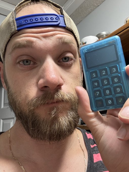 A man with a beard wearing a backwards cap holds a blue translucent payment terminal up to the camera.