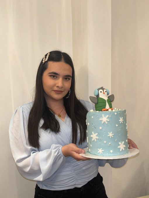 A young woman with long dark hair wearing a light blue blouse and pearl hair accessory, holding a light blue winter-themed cake decorated with snowflakes and a penguin figurine on top.