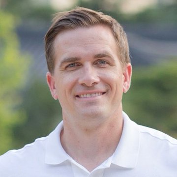 A headshot of a smiling man with short brown hair wearing a white polo shirt, photographed outdoors with greenery in the background.