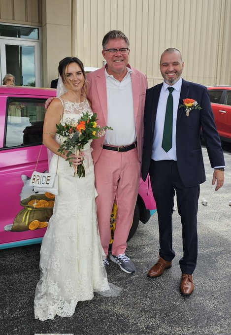 A bride in a white lace gown holding an orange bouquet stands between a man in a pink suit and a groom in a navy suit with a green tie, posing in front of a pink car decorated with a "BRIDE" sign featuring cartoon gold coins — fitting the #UFDwedding theme of a marriage celebration within the crypto community.
