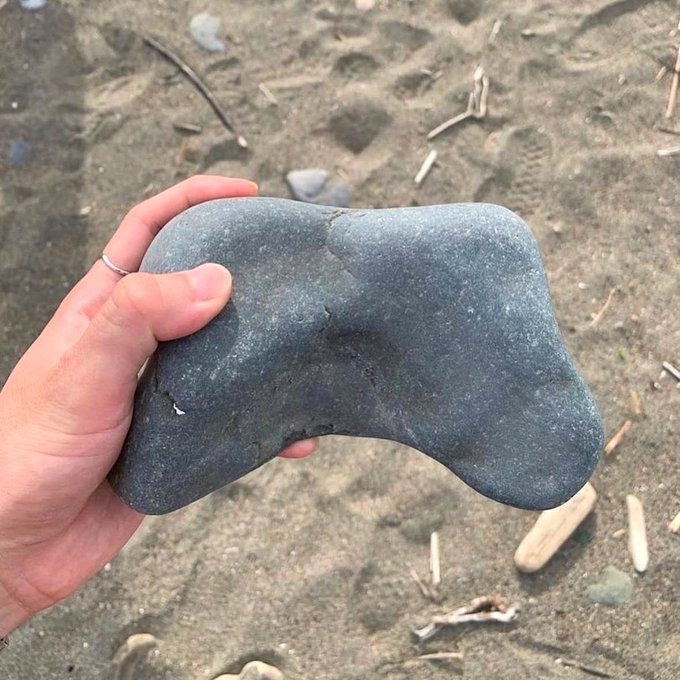 A hand holding a smooth gray rock on a sandy beach, with the rock's unusual wavy shape resembling a video game controller.