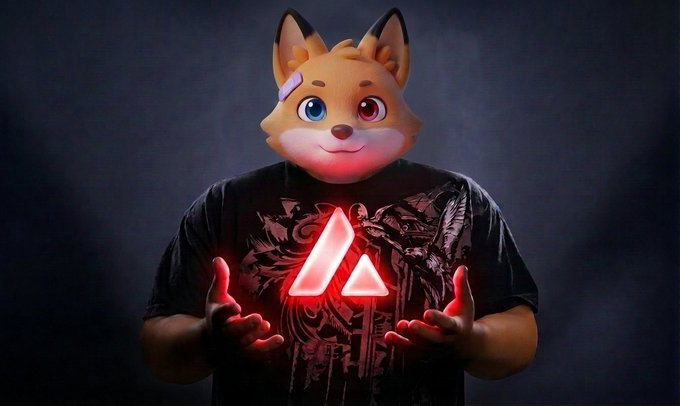 A person wearing a black graphic t-shirt featuring a glowing red geometric logo stands against a dark background while wearing an orange fox-like mascot head with large blue and red eyes.