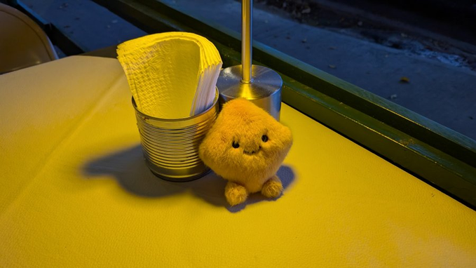 A small plush yellow creature sits beside a tin can holding folded yellow paper napkins on a yellow table near a window.