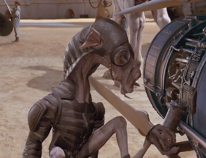 A computer-generated sci-fi scene shows a pale, armored humanoid creature crouched beside a large cylindrical machine in a sandy arena.