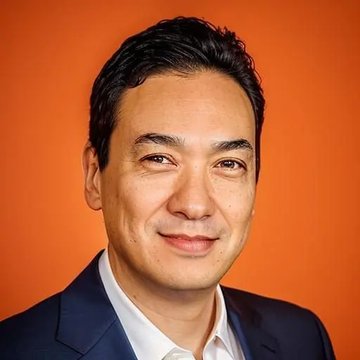 A man in a navy suit and white shirt smiles against a vibrant orange background, presumably Simon Gerovich, CEO of Metaplanet, who recently commented on the emergence of the first Asian Bitcoin cycle.