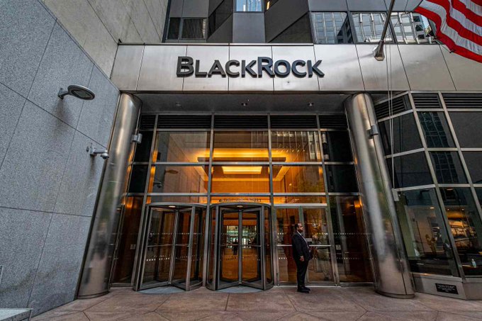 The image shows the entrance to BlackRock's headquarters, featuring its name displayed prominently above glass revolving doors, with a man in a mask standing outside and an American flag visible. This visual ties directly to the post, as BlackRock—the world's largest asset manager—has been actively purchasing Bitcoin through its ETFs, with the caption highlighting their recent $244.42 million in BTC acquisitions signaling institutional re-entry into the crypto market.