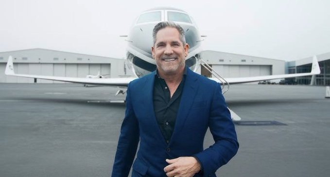 A smiling man in a blue blazer and black shirt stands on an airport tarmac with a private jet and hangars in the background.