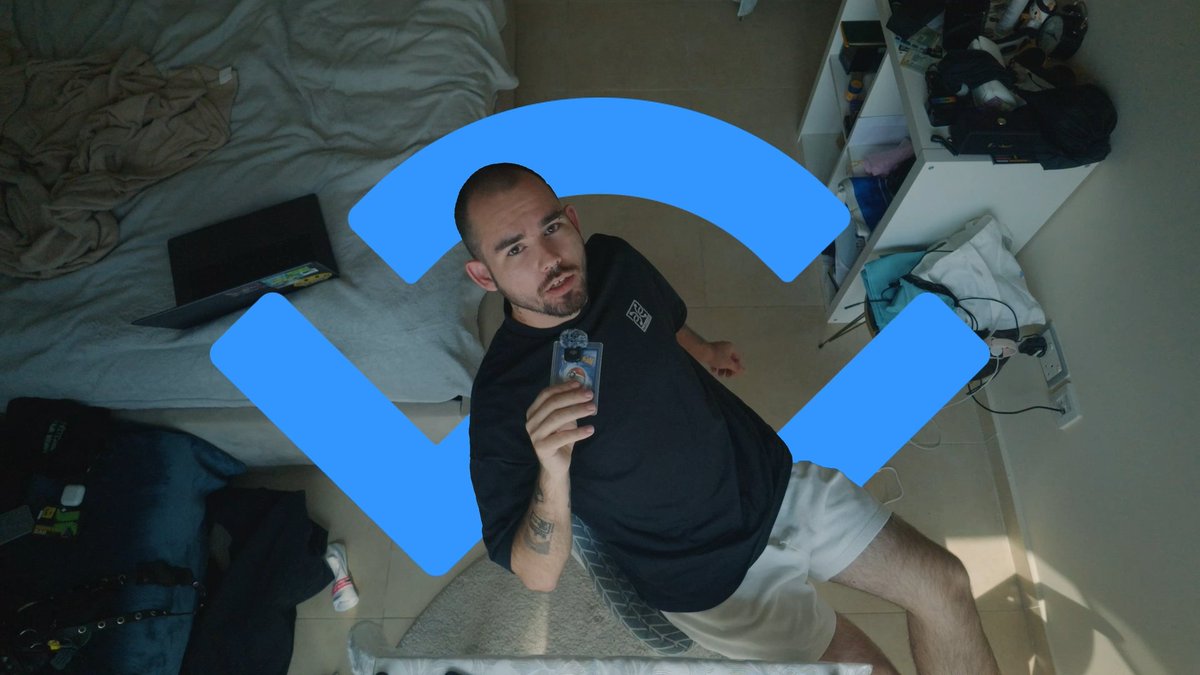 A man sits in a bedroom holding a small card or package, with a large blue geometric logo overlaid on the image; the room contains a bed with rumpled sheets, a laptop, and scattered belongings.