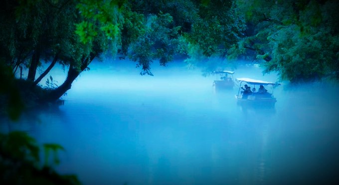 Two small boats with canopies glide through a misty, blue-tinted river surrounded by lush green trees, creating a serene and dreamlike atmosphere that captures the "time to recharge" message of the post.