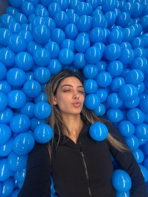 A woman lying in a ball pit filled with bright blue plastic balls, posing with her eyes closed and lips puckered.