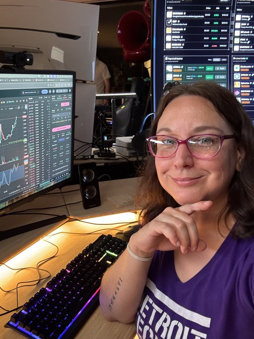 A woman with pink glasses and a purple "Detroit" jersey smiles thoughtfully at her desk, surrounded by crypto trading screens showing various tickers like SOL and pump.fun metrics, capturing the quiet resilience of someone still holding their position in the crypto space.