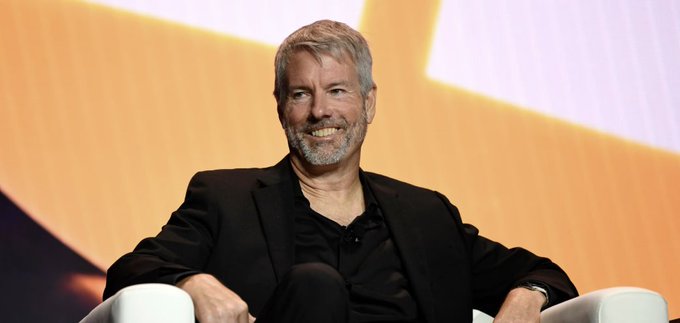 An older man with white hair and beard, wearing a black blazer, smiling while seated in a white chair against a warm yellow and orange backdrop.