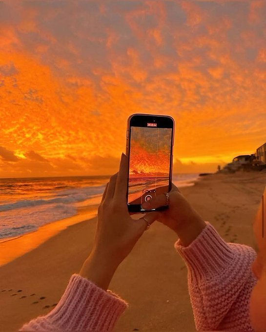 The image shows a person taking a picture of themselves with their cell phone at the beach during sunset. The sky has an orange hue, creating a beautiful backdrop for the photo. The person holding the phone appears to be capturing this moment as part of their daily life or documenting their experience on social media platforms like Instagram.