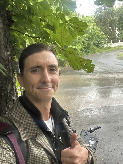The image features a man wearing a brown jacket standing under a tree with his thumb up in a thumbs-up gesture. The man appears to be smiling as he poses for the picture. The background of the photo shows a street, suggesting that they are in an urban setting or near a road intersection. There is also a bicycle visible in the image, which might indicate that the man has recently ridden his bike and is now posing with it.