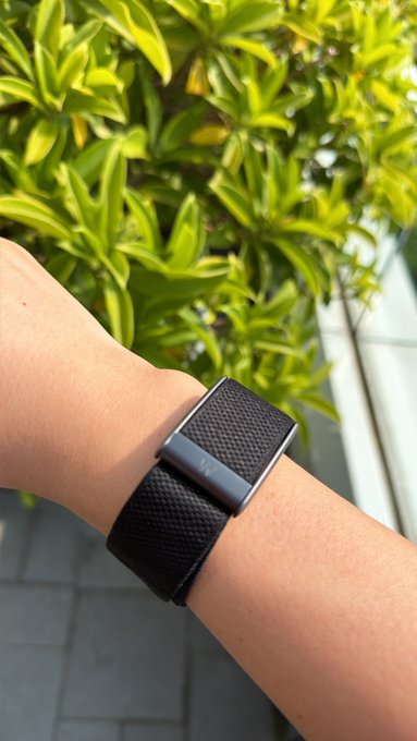A close-up of a wrist wearing a black WHOOP fitness band with a textured strap and metal clasp, set against a backdrop of vibrant green foliage and tiled flooring, illustrating the post's announcement of getting a WHOOP device.