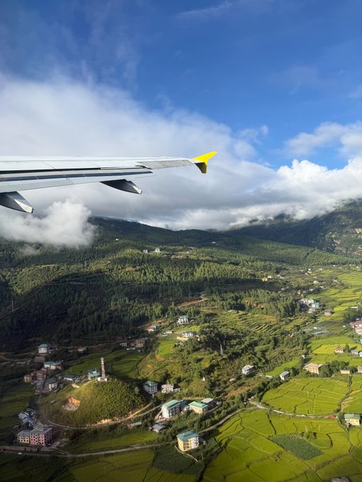 An aerial view from an airplane window reveals lush green rice terraces and small mountain villages in what appears to be Bhutan, with the plane's wing stretching across the cloudy sky above—symbolizing a scenic aerial perspective on Robinhood Chain, which, though currently overshadowed, is taking flight with emerging innovations and poised for growth.