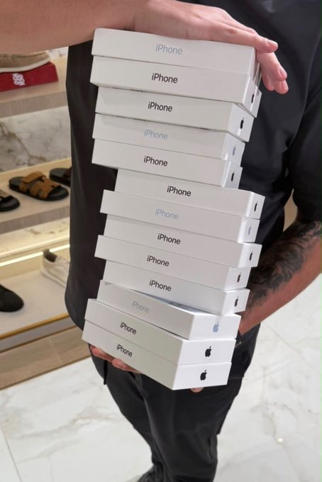 A stack of 11 white iPhone boxes being carried by a person, tied to a fake "iPhone 18 Pro Max Giveaway" post claiming participants can pick a number from 20–72 to win — a clear giveaway scam using the iPhone imagery to lure engagement.