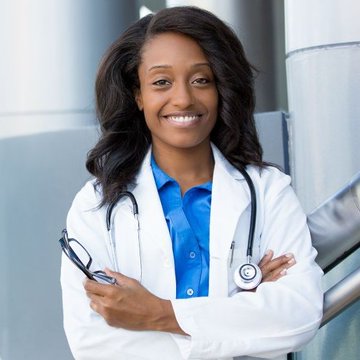 A smiling Black woman wearing a white doctor's coat over a blue button-up shirt stands with her arms crossed, holding a stethoscope.