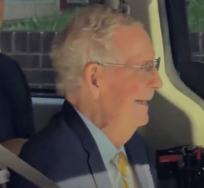 A close-up profile shot of an elderly man with white hair and glasses, wearing a blue suit and a yellow patterned tie, seated in a car with a seatbelt visible.