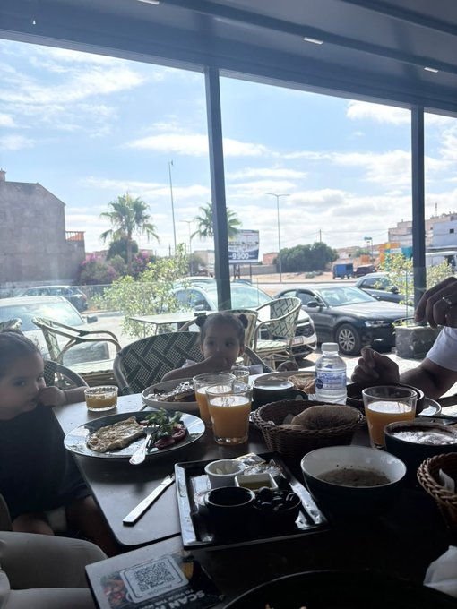 A family enjoys breakfast at a restaurant table with large windows overlooking a parking lot and palm trees, with plates of food, juice, and coffee on the table.