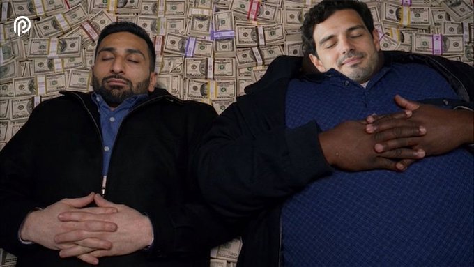 The image shows two men lying down next to each other with their arms wrapped around one another, both appearing to be sleeping or relaxed. They are positioned against a backdrop of dollar bills scattered across the area, creating an unconventional setting for this scene. The caption provided includes information about a popular meme involving these men and money.
