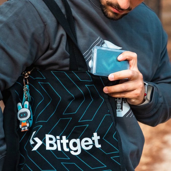 A close-up of a person holding a deck of cards while wearing a black messenger bag with teal geometric patterns, branded with the Bitget logo and featuring a small robot character keychain.
