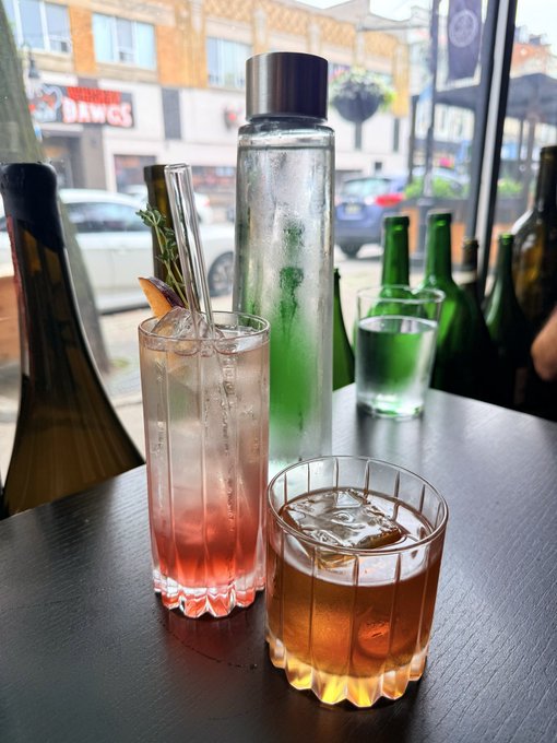 Two cocktails—a pink-hued spritz garnished with rosemary and an orange-hued drink on the rocks—sit on a bar table alongside a tall bottle of water, with a "Dawgs" storefront visible through the window, perfectly capturing the relaxed Friday date night vibe away from the charts and crypto work.