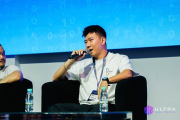 A man speaks into a microphone while seated on stage at a conference, with an "Ultra" logo visible in the corner.