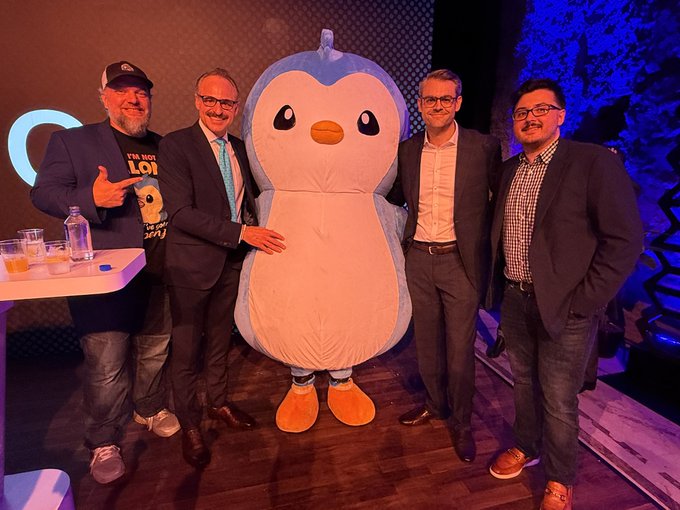 Four men in business and casual attire pose for a photo with a large blue and white penguin mascot costume at what appears to be an indoor event or party venue.