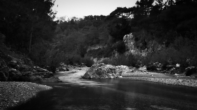 A black and white landscape photograph of a calm river flowing through a rocky valley surrounded by dense trees.