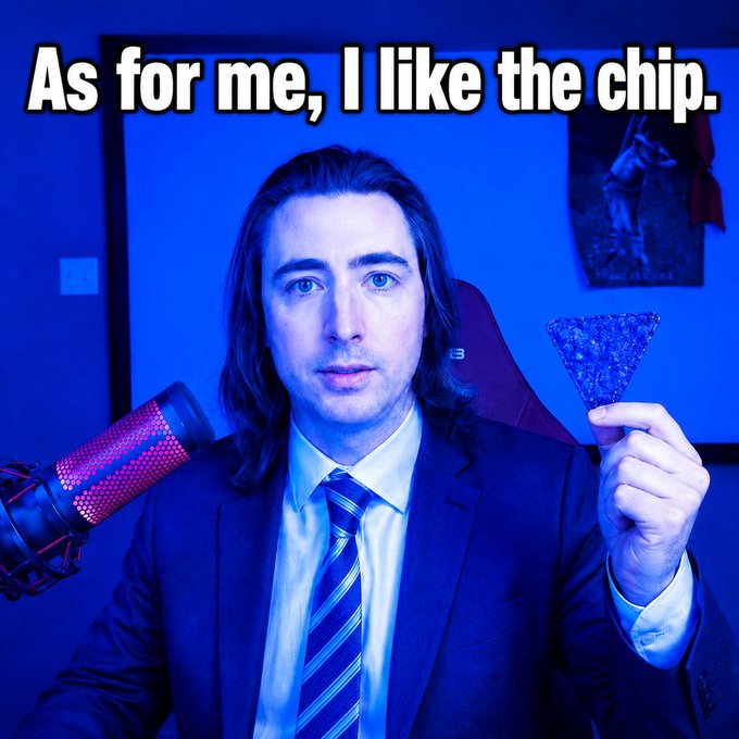 A man with long dark hair wearing a suit and striped tie, sitting in front of a red-lit microphone, holding up a triangular tortilla chip against a blue-lit background, with the caption "As for me, I like the chip."
